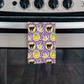 A halloween kitchen towel with a purple background and a watercolor pattern of slice and bake cookies for halloween surrounded by sprinkles. Includes ghost cookies, black cat cookies, and jack o lantern cookies. Displayed hanging on oven handle. halloween kitchen towel, halloween kitchen towels, halloween tea towel, halloween tea towels, kitchen towels for halloween, halloween kitchen hand towels
